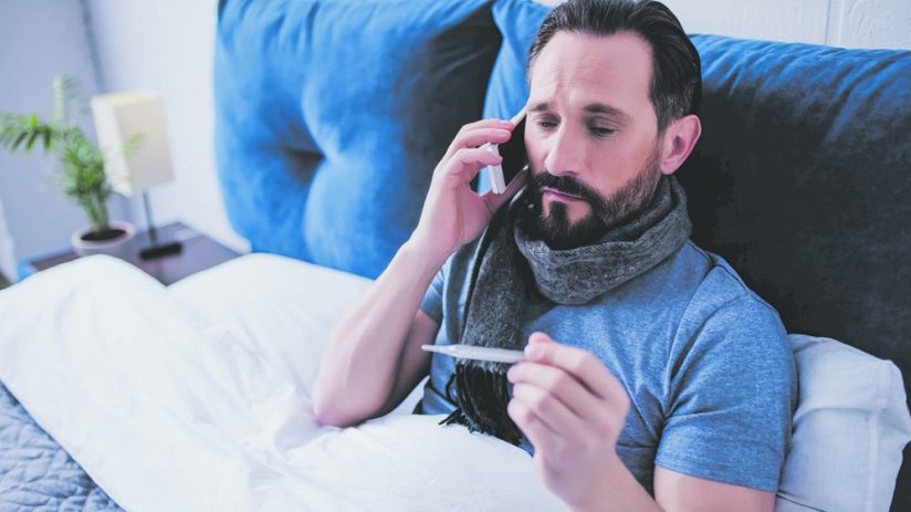 portrait of sick man holding thermometer and calling a doctor while sitting on the bed