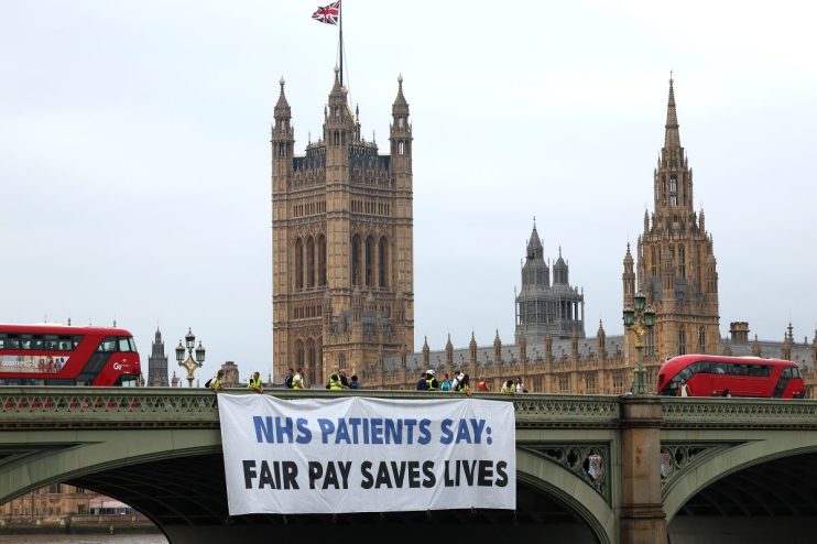 LONDON, ENGLAND - JUNE 27: Just Treatment, a protest group, unfurl a banner in support of striking young doctors, on Westmimnster Bridge, on day one of the British Medical Association (BMA) strike action on June 27, 2024 in London, United Kingdom. Junior doctors, who are members of the BMA, are striking in the final week before Britain goes to the polls. Despite receiving an 8.8% pay rise last summer and an additional 3% offer, the BMA rejected the 3% offer as it doesn't compensate for the over 25% loss in real-term pay over the past 15 years. The BMA is demanding full pay restoration to reverse these real-term cuts since 2008-09, along with a new pay mechanism to prevent future pay decreases due to inflation and the cost of living. (Photo by Peter Nicholls/Getty Images)