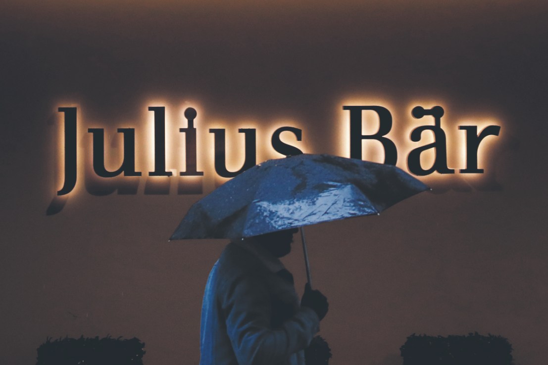 A pedestrian sheltering under an umbrella passes a Julius Baer Group Ltd. branch in Zurich (Photographer: Stefan Wermuth/Bloomberg via Getty Images)